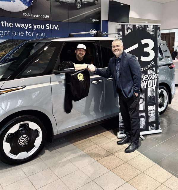 A handshake through the driver’s window of a silver Volkswagen ID. Buzz beside a Less Than 3 banner in a Volkswagen showroom