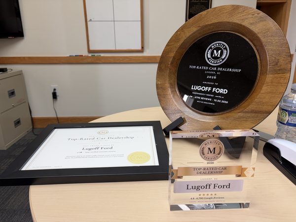 A round wooden plaque, a clear acrylic desk award and a framed certificate on a table, each naming Lugoff Ford a 2026 Meritly Verified Top-Rated Car Dealership in Lugoff, SC